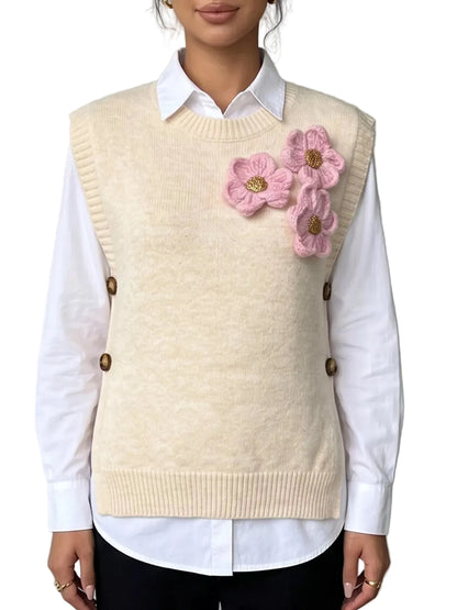A cream-colored knit sweater vest worn over a white collared shirt. The vest features decorative pink crocheted flowers on the left chest and brown buttons on the sides, styled with black pants against a neutral gray background.