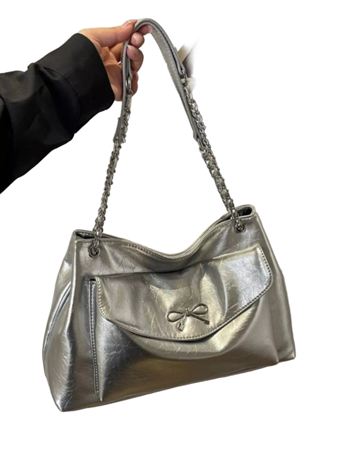 A metallic silver shoulder bag with a chain strap, held by a hand wearing a black sleeve. The bag features a front flap with a bow detail and a zippered pocket.