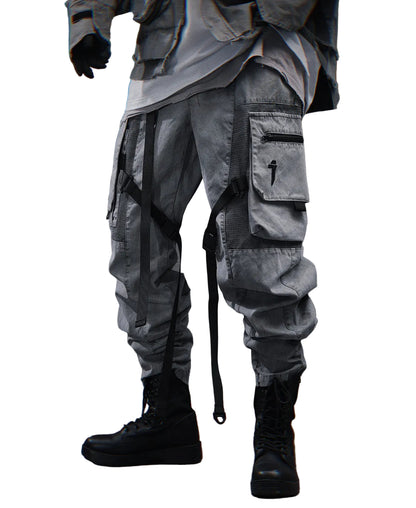A person wearing loose, oversized cargo-style pants in a washed gray tone, styled with black combat boots and dark gloves, standing against a textured gray wall.