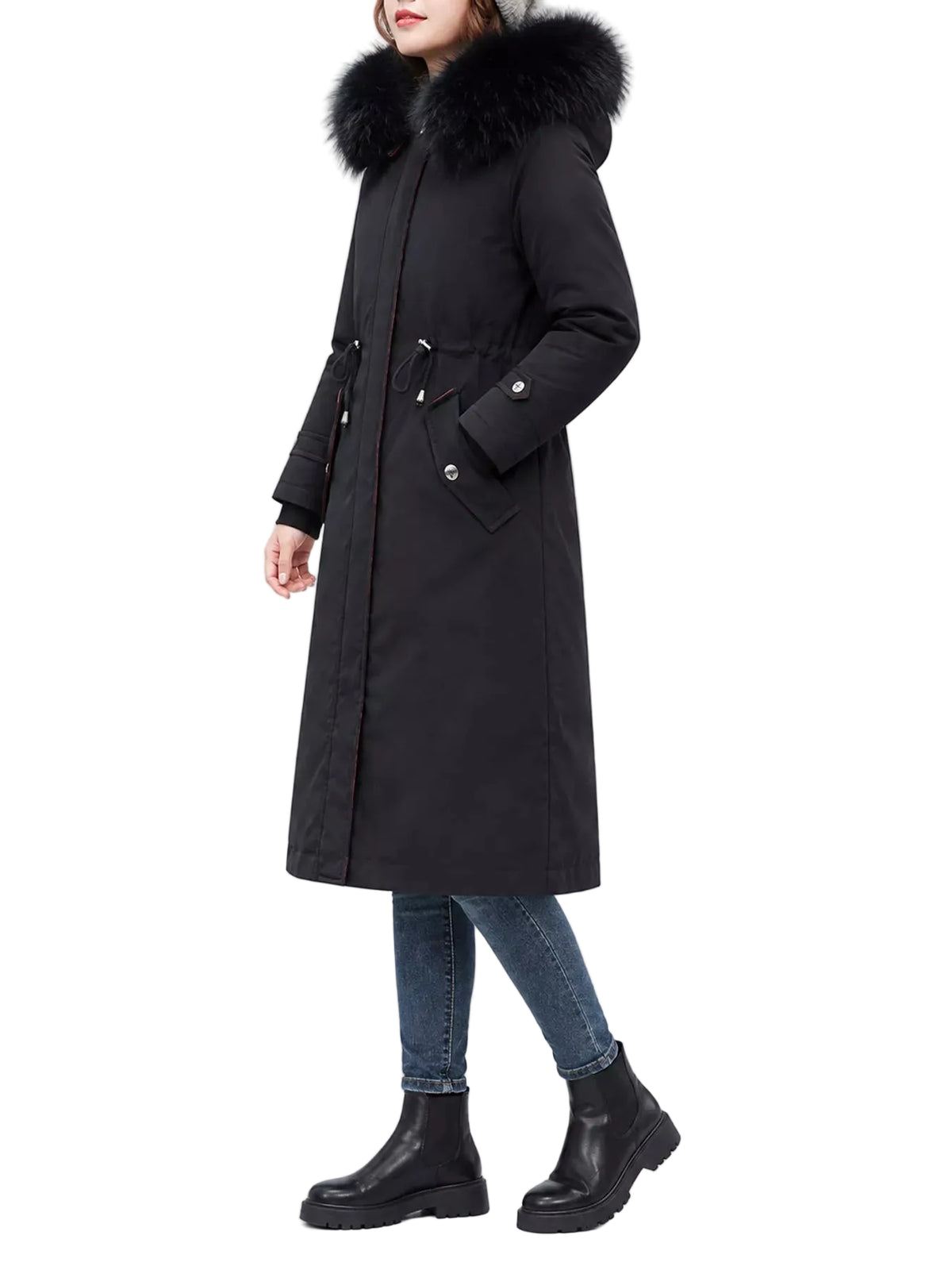 A woman wearing a long black parka coat with a large black fur-trimmed hood, paired with blue jeans and black ankle boots, photographed against a plain light gray studio background.