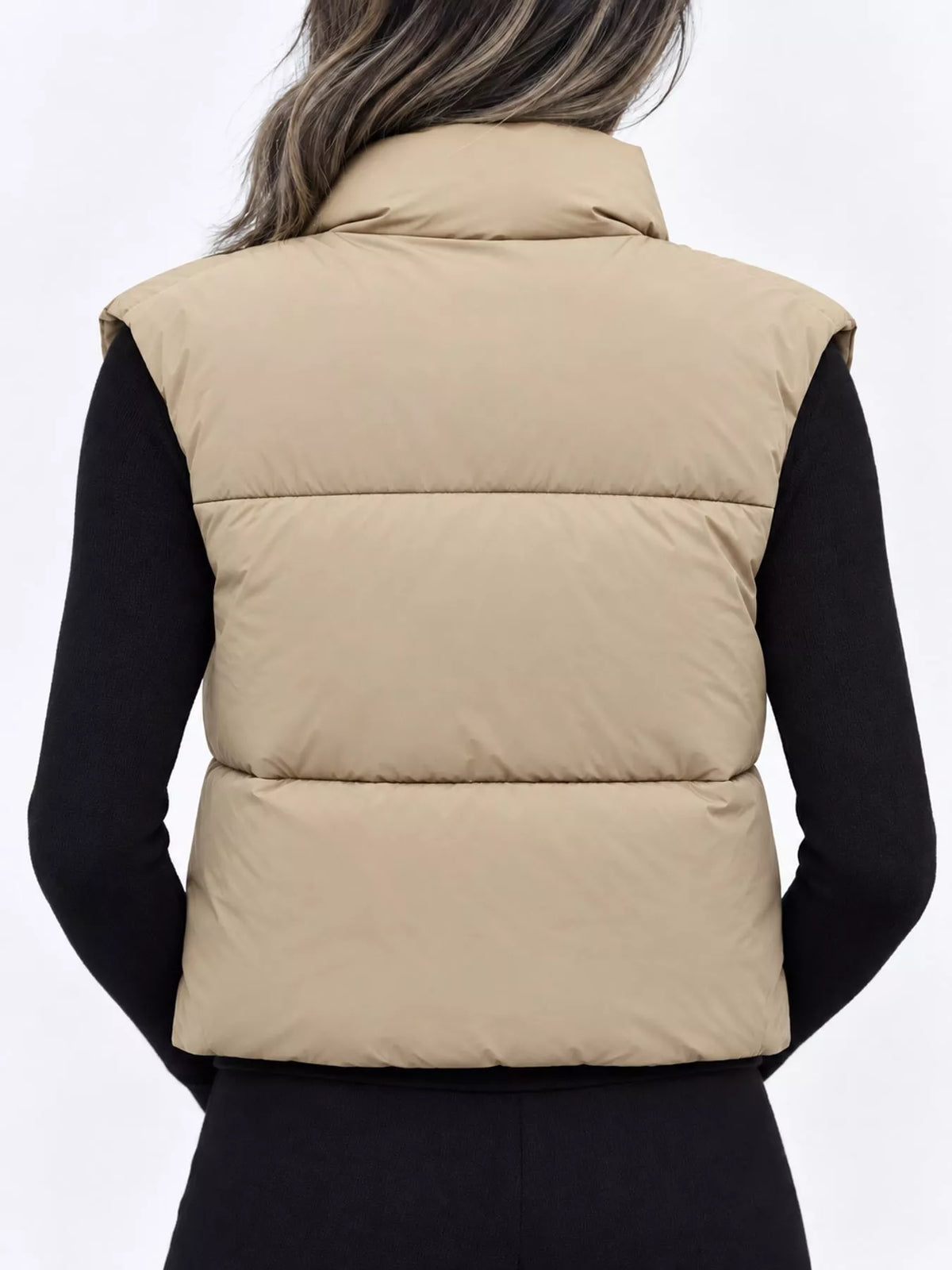 A person shown from the back wearing a sleeveless puffer vest in a light tan/beige color over a black long-sleeve top, against a plain light gray studio background.
