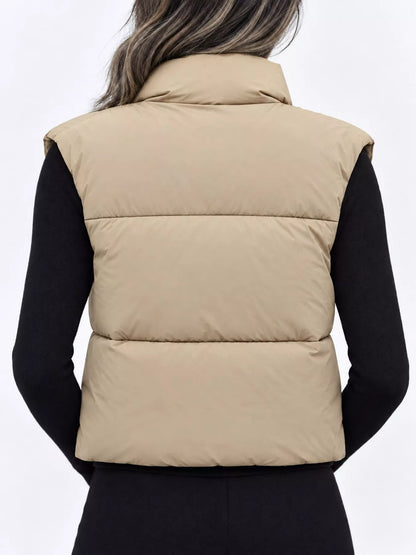 A person shown from the back wearing a sleeveless puffer vest in a light tan/beige color over a black long-sleeve top, against a plain light gray studio background.
