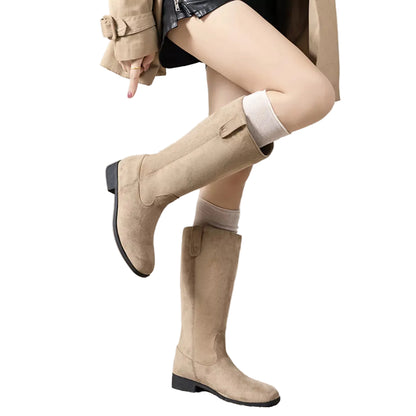 A person wearing knee-high beige suede boots with a low block heel, paired with white socks and a beige trench coat, standing on a textured rug in a minimalist indoor setting.
