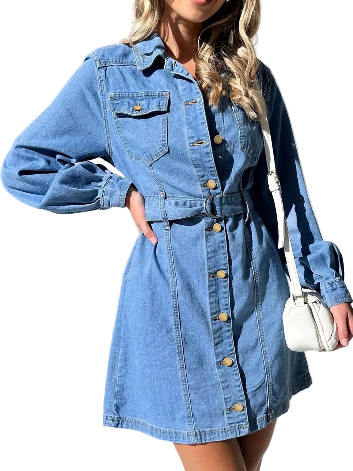 A woman wearing a light blue denim long-sleeve shirt dress with a button-down front, chest flap pockets, and a matching denim belt. She is holding a small white shoulder bag against a background of white draped curtains.