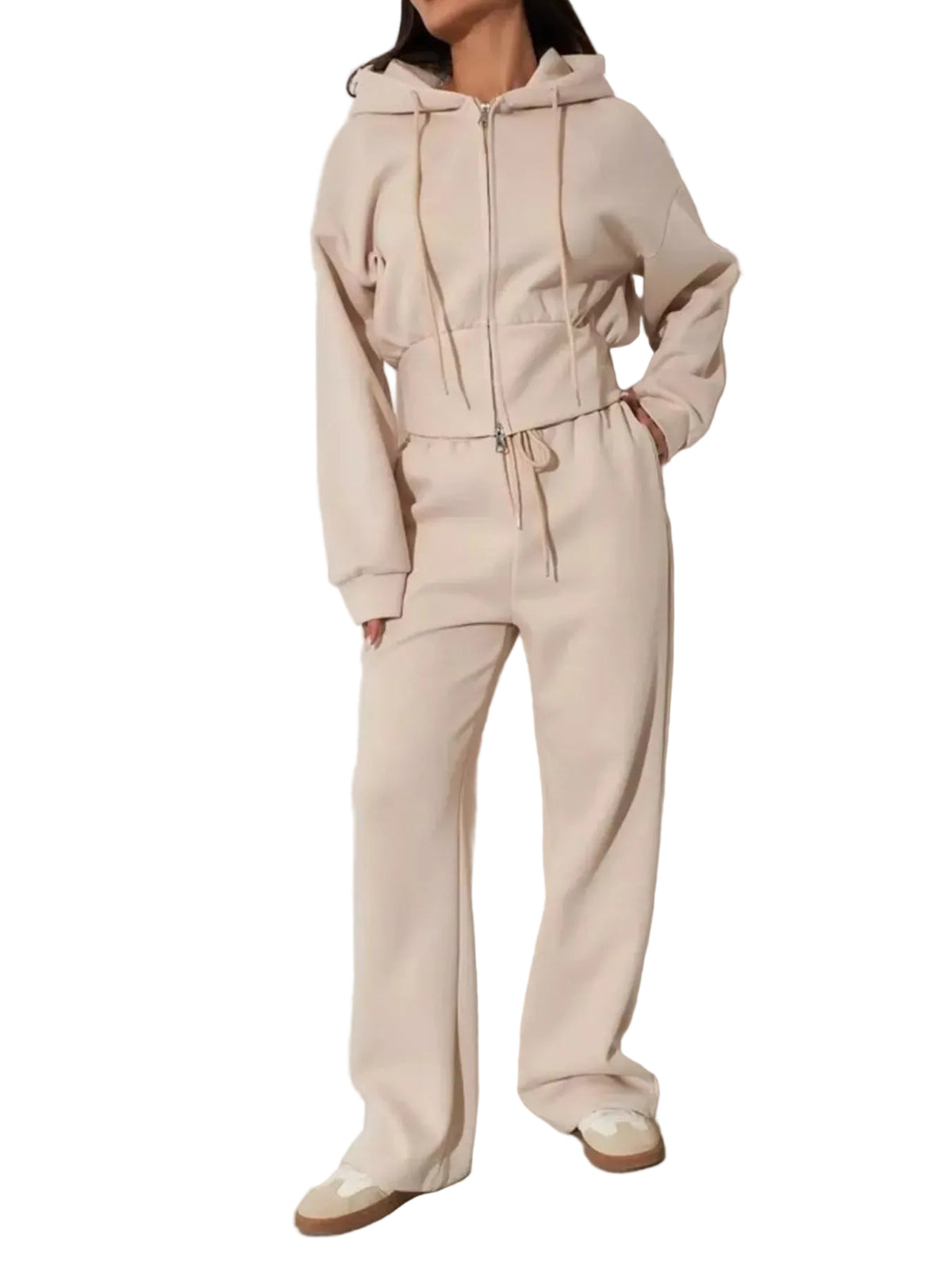 A model wearing a light beige two-piece tracksuit featuring a cropped zip-up hoodie and matching wide-leg sweatpants, standing against a minimalist off-white wall.