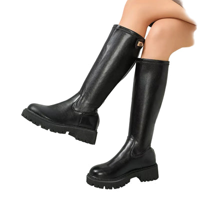 A person wearing black knee-high leather boots with chunky lug soles, shown in a bright modern interior with a white bathtub and glass floor panels.