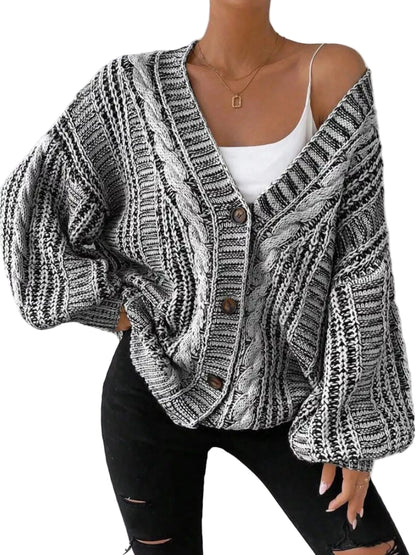 A woman wearing a chunky knit cardigan with a black and white cable pattern, styled over a white camisole and black ripped jeans against a textured white wall.