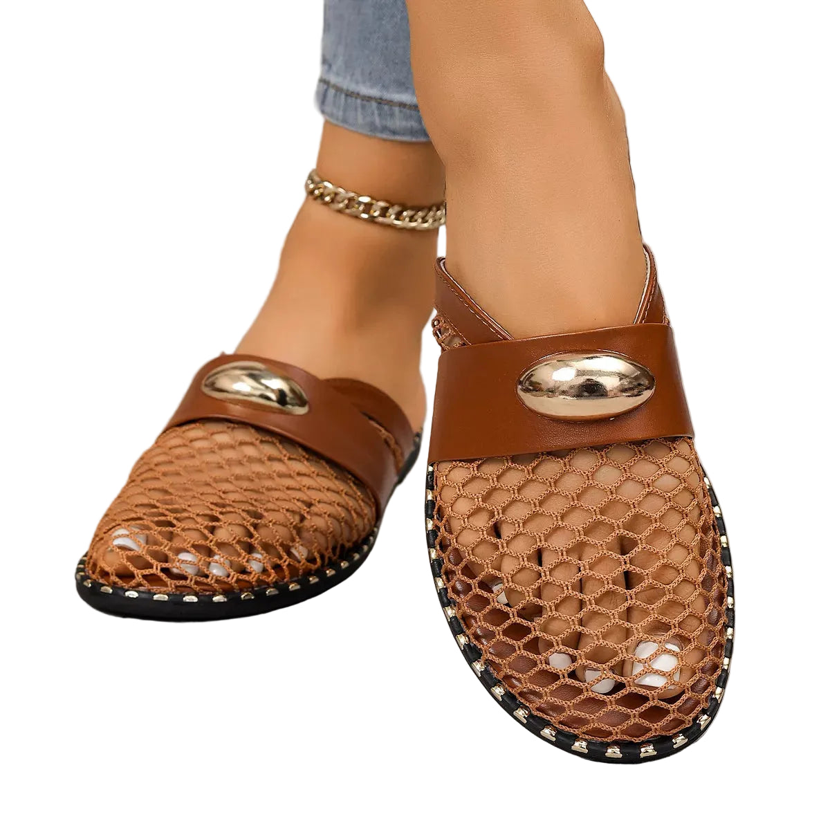A pair of women's brown mesh mule sandals with a wide brown strap and shiny gold oval hardware, shown on a model's feet against a gray concrete floor.