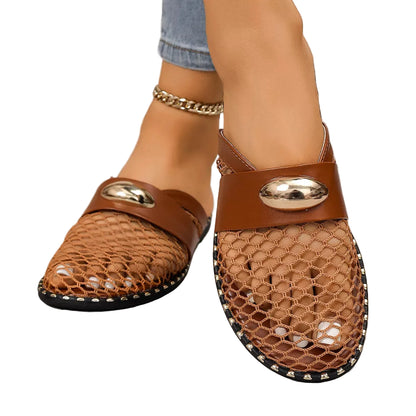 A pair of women's brown mesh mule sandals with a wide brown strap and shiny gold oval hardware, shown on a model's feet against a gray concrete floor.