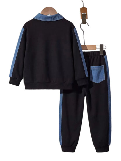 A two-piece kids' tracksuit set consisting of a black long-sleeve top with light blue contrast panels on the collar and sleeves, and matching black pants with light blue side stripes and a blue back pocket, displayed on wooden hangers against a gray wall with books in the background.