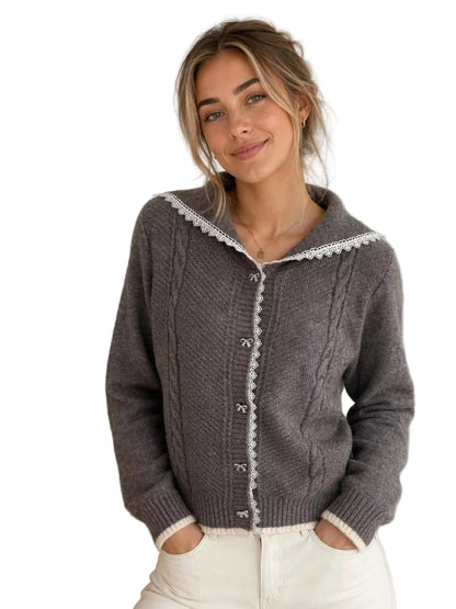 A woman wearing a dark gray cable-knit cardigan with white lace trim and white buttons, paired with white pants, standing in a brightly lit room.
