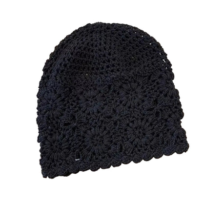 A black crocheted beanie hat with a textured floral pattern and scalloped edge, laid flat on a light wood surface with a cream fabric and striped cloth in the background.