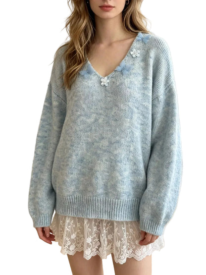 A woman wearing a light blue marled knit sweater with decorative white and blue flower embellishments on the neckline, paired with a white lace skirt in a bright indoor room.