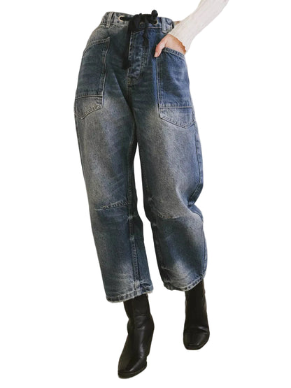 A person wearing loose-fit blue denim jeans with a black drawstring waist, paired with a white top and black ankle boots, standing against a plain light wall.