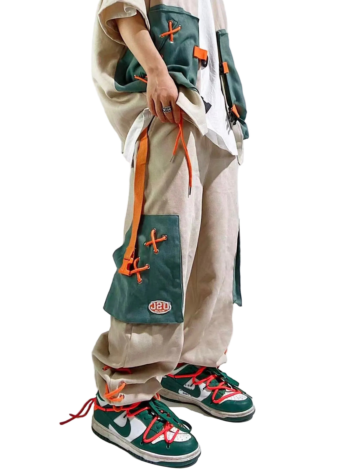 A person wearing loose beige cargo-style pants with large green patch pockets and orange laces, a white shirt, and green sneakers with orange laces, photographed against a light gray studio background.