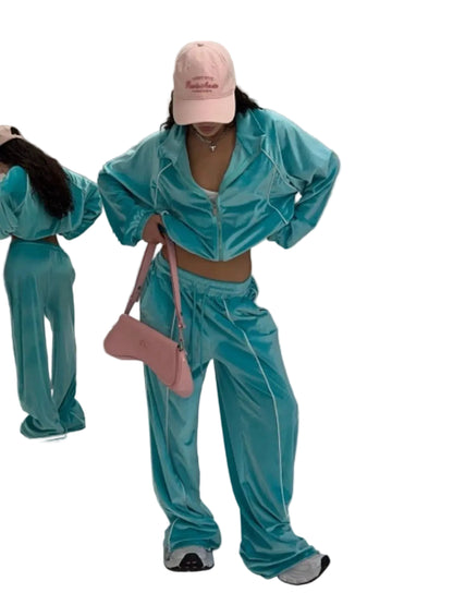 A person modeling a matching turquoise-blue velour tracksuit with white piping, a pink baseball cap, and a pink shoulder bag in a studio setting.