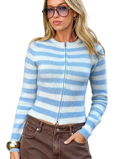 A woman wearing a light blue and white striped zip-up knit sweater, brown pants, and sunglasses, posing outdoors against a light blue wall with green plants on the left.