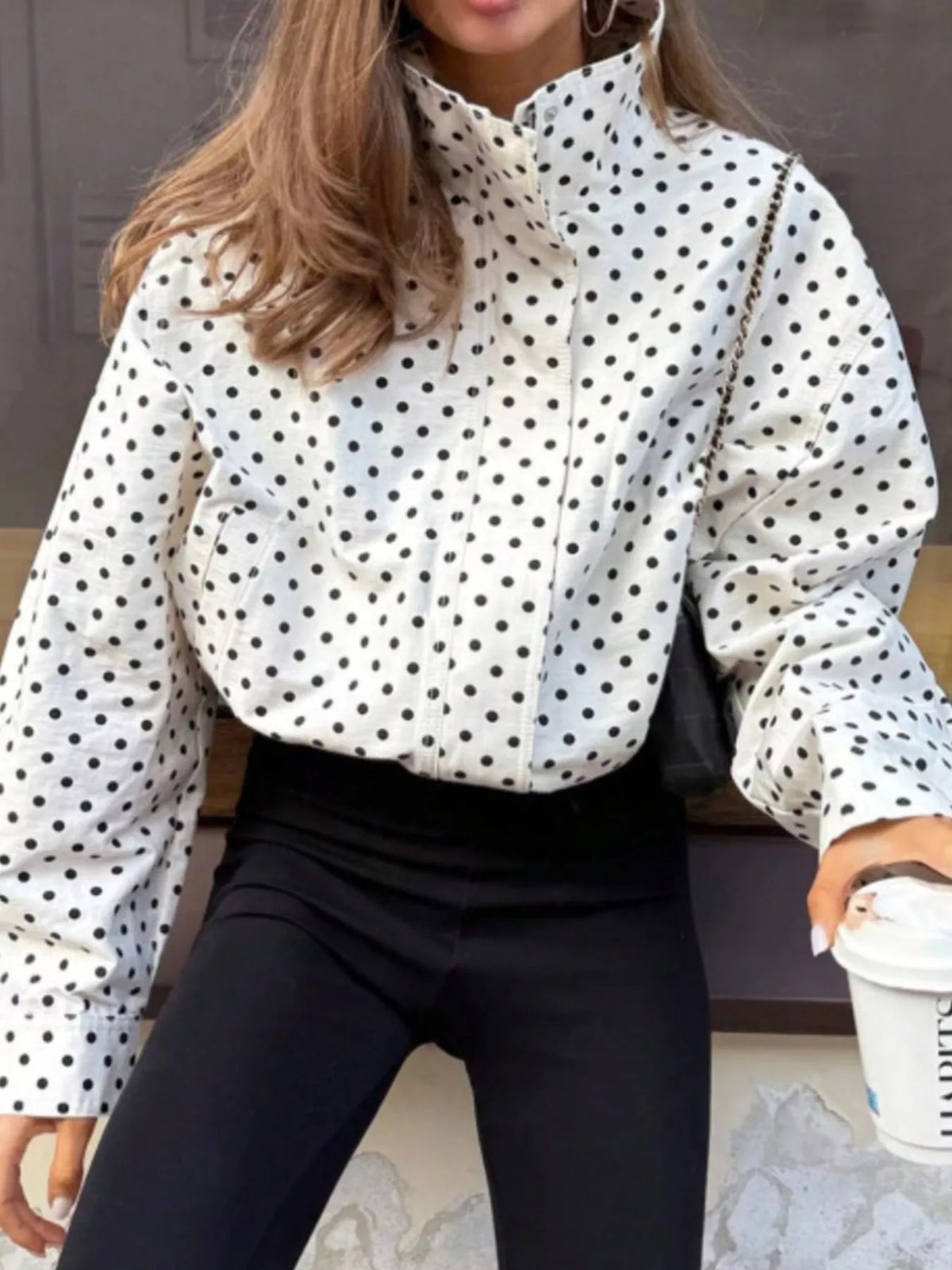 A woman wearing a white long-sleeve blouse with black polka dots and black pants, holding a white coffee cup.