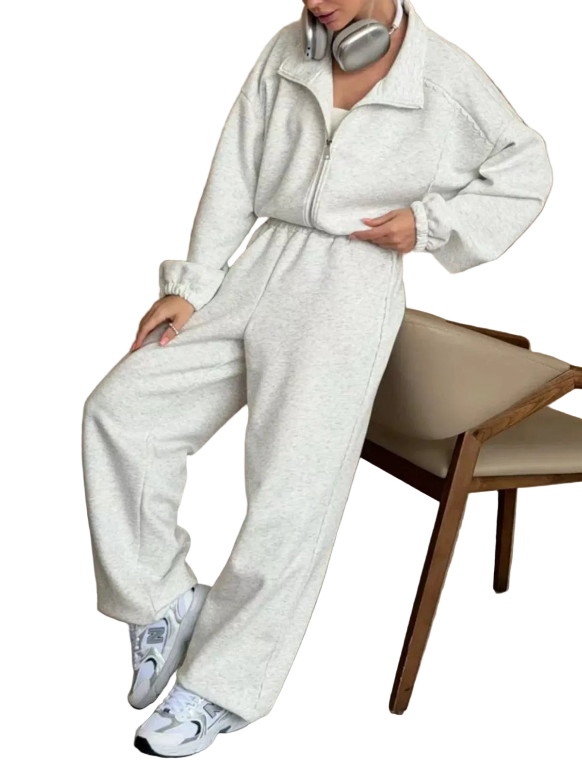 A person wearing a light heather gray zip-up sweatshirt and matching wide-leg sweatpants, seated on a beige upholstered wooden chair against a wood-paneled wall. Silver headphones are around the neck, and white sneakers are visible.