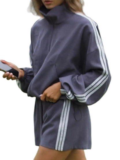 A woman wearing an oversized dark gray athletic tracksuit with white stripes down the sleeves and side, standing on a green tennis court.