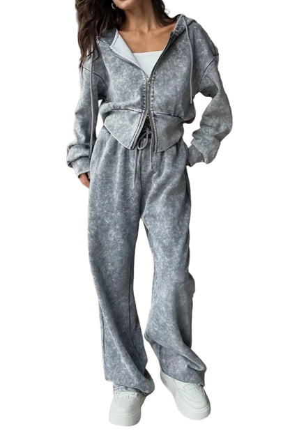 A woman models a matching gray zip-up hoodie and wide-leg sweatpants set with a mottled or marbled texture, styled with white sneakers in a modern living room setting.