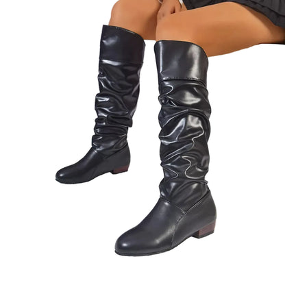 A pair of black knee-high slouchy boots with a low block heel, shown on a model seated on a white bench in a snowy outdoor-themed setting.