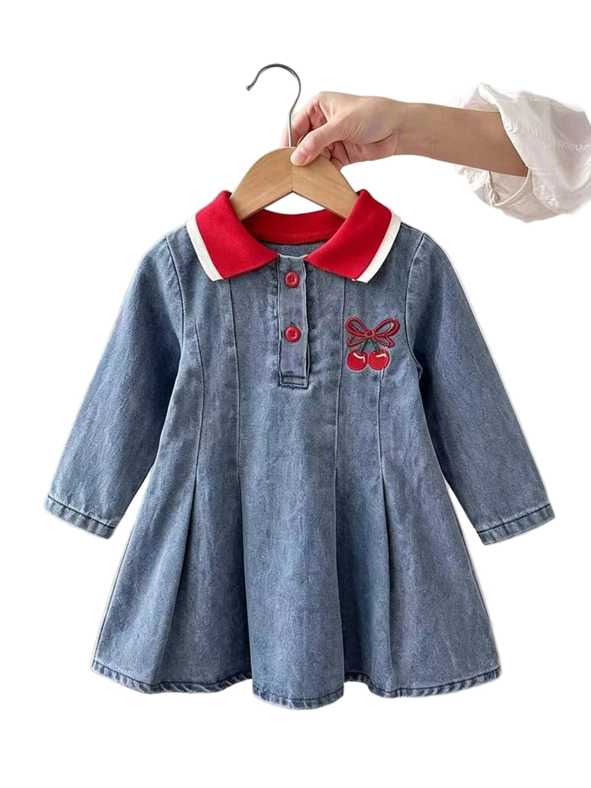 A toddler girl's blue denim dress with a red and white collar, red buttons, and a red bow embroidery on the chest, held on a wooden hanger by a person wearing a white dress.