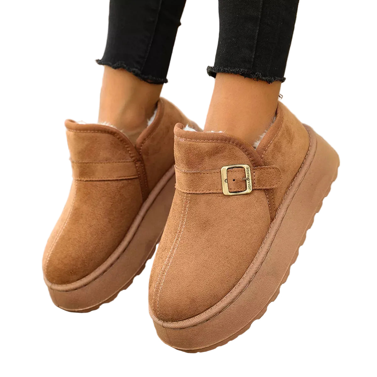 A pair of women's platform slip-on boots in tan suede with a matching strap and gold buckle, shown being worn on a wooden deck.