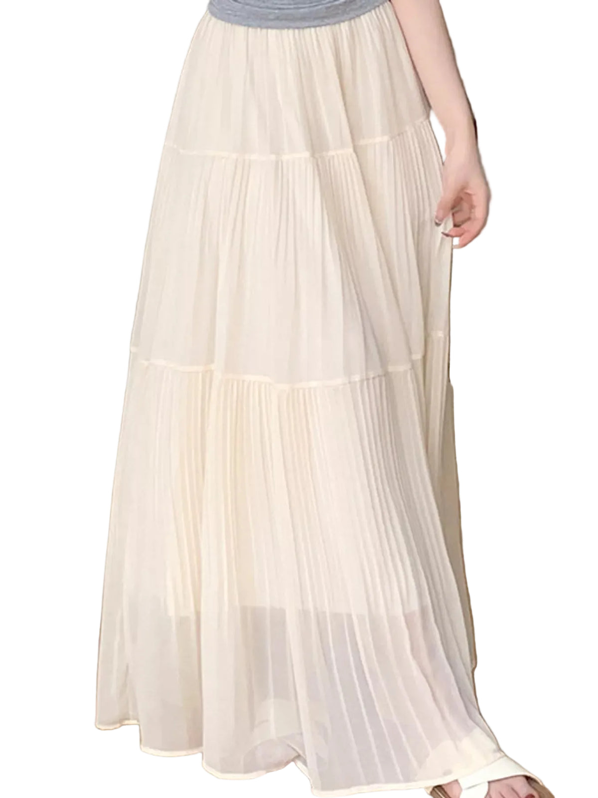 A person wearing a long, tiered pleated maxi skirt in a light cream or off-white color, paired with a gray top and white sandals, standing on a light wood floor.