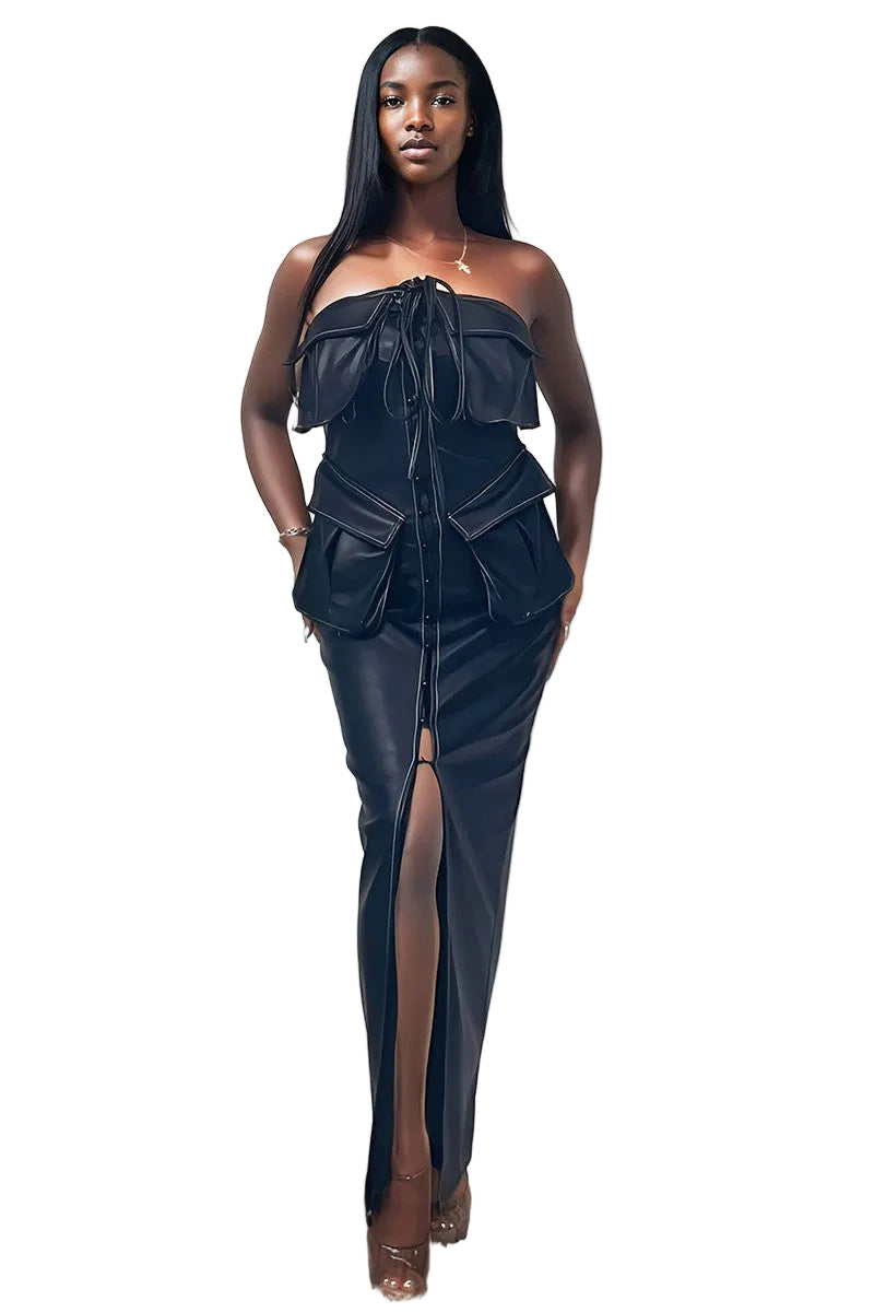 A woman modeling a black strapless faux-leather dress with a structured corset-style bodice, ruffled shoulder details, side pockets, and a high front slit, standing against a plain off-white wall.