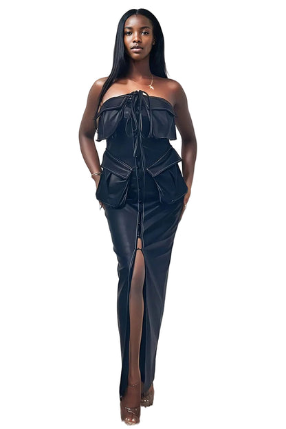 A woman modeling a black strapless faux-leather dress with a structured corset-style bodice, ruffled shoulder details, side pockets, and a high front slit, standing against a plain off-white wall.