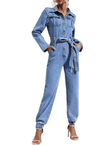 A woman models a light blue denim one-piece jumpsuit with a button-up front, chest pockets, a tie waist belt, and cuffed ankles, paired with white heeled sandals against a white wall with palm leaf shadows.