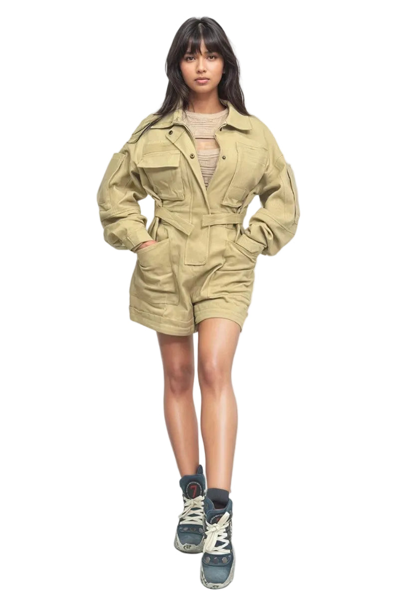 A model stands against a light gray studio background wearing a beige utility jumpsuit with a belt, layered over a beige ribbed top, paired with blue and gray sneakers.