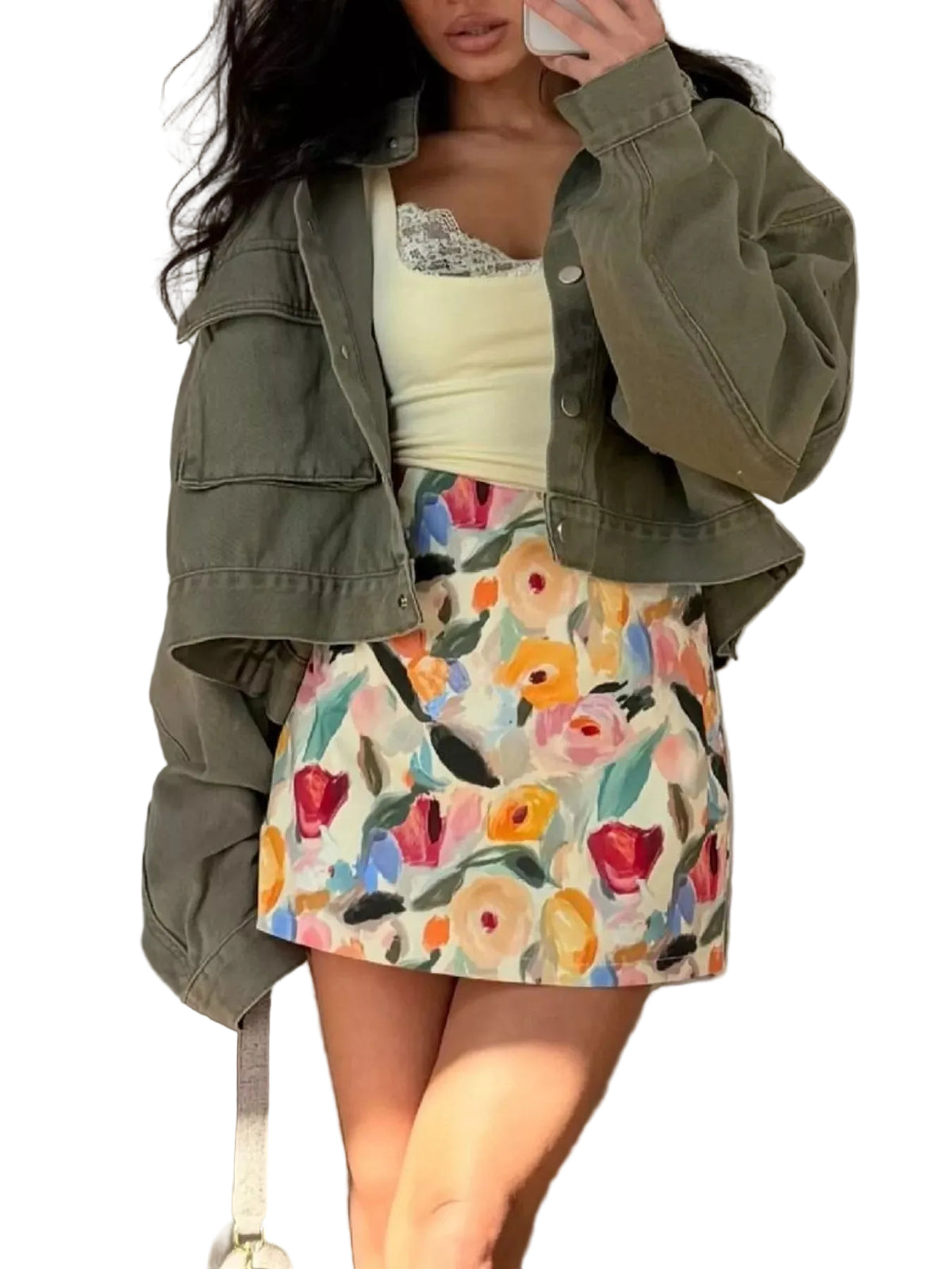 A woman wearing a cropped olive green denim jacket over a pale yellow top and a colorful floral mini skirt, posing in front of a neutral curtain backdrop.