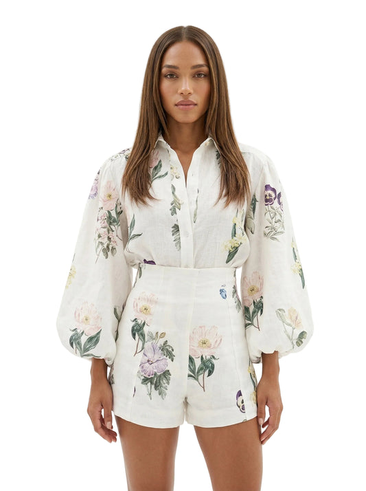 A woman modeling a matching white two-piece set with a delicate floral print, featuring a long-sleeve button-up shirt and coordinating shorts, set against a plain white studio background.