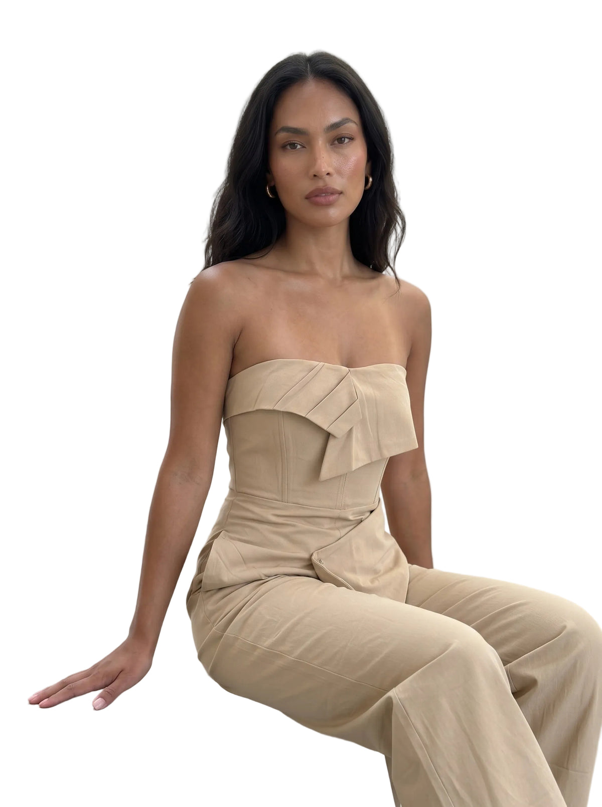 A woman sits on a concrete ledge wearing a beige strapless jumpsuit with a structured ruffled bodice and wide-leg pants, posed against a light concrete wall.