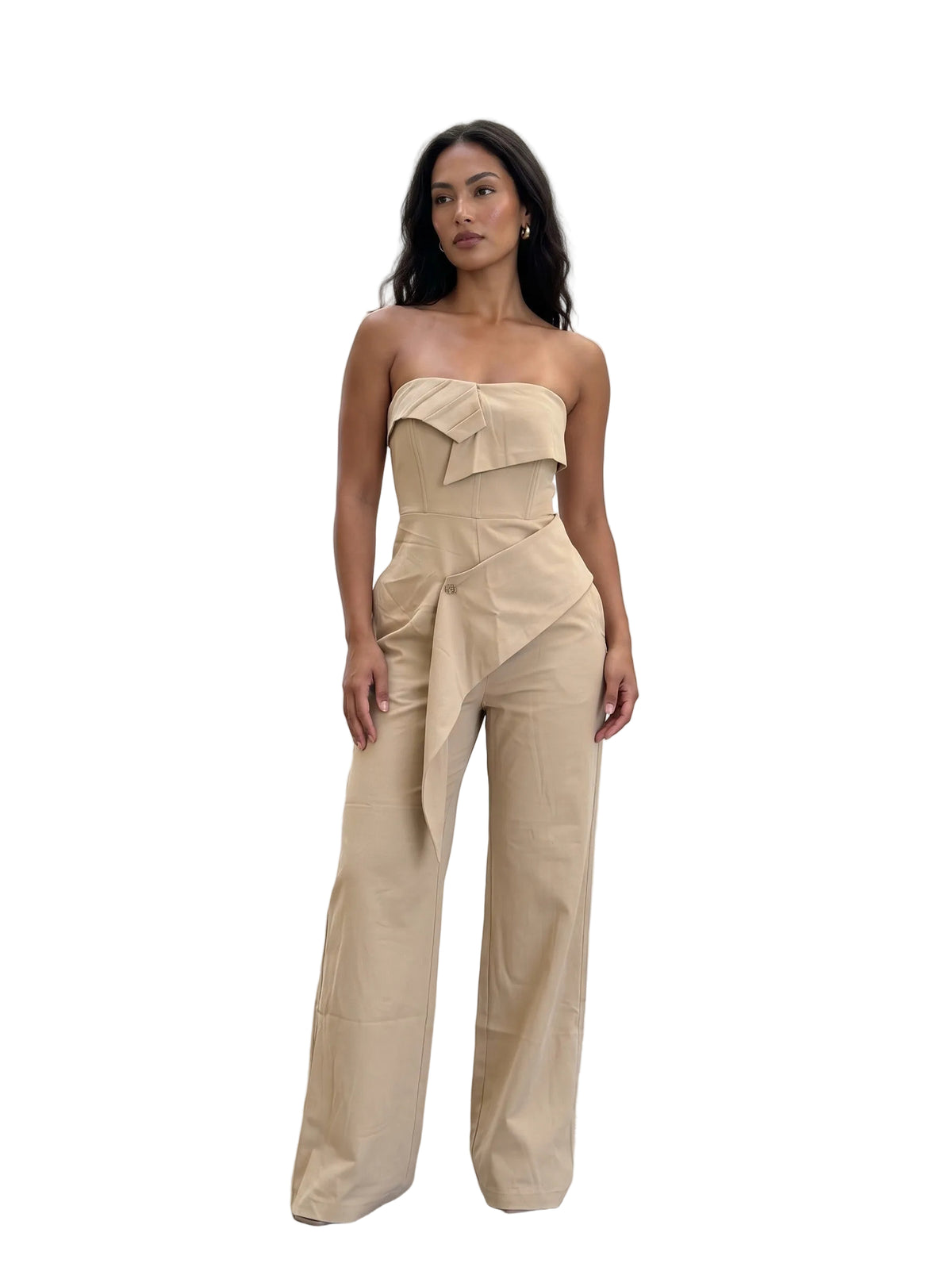 A woman stands against a light concrete wall wearing a beige strapless jumpsuit with a structured ruffled bodice and wide legs.