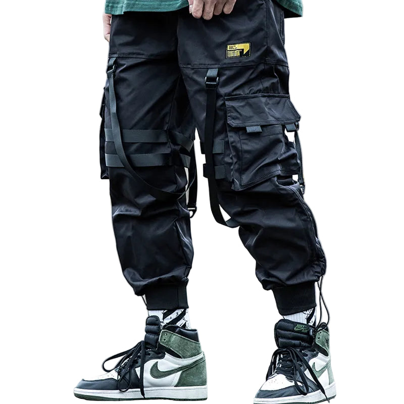 Black cargo jogger pants with multiple utility pockets, compression straps, and elastic cuffs, shown on a model wearing high-top sneakers on an outdoor street.
