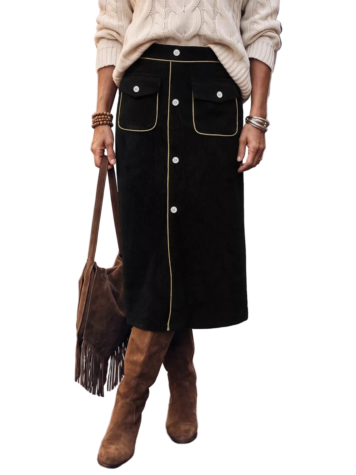 A person standing on a city street wearing a black midi skirt with gold contrast piping and front buttons, a cream cable-knit sweater, and brown suede knee-high boots, while carrying a brown fringe shoulder bag.