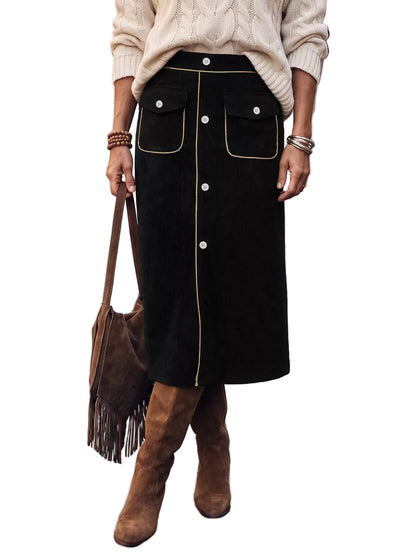 A person standing on a city street wearing a black midi skirt with gold contrast piping and front buttons, a cream cable-knit sweater, and brown suede knee-high boots, while carrying a brown fringe shoulder bag.