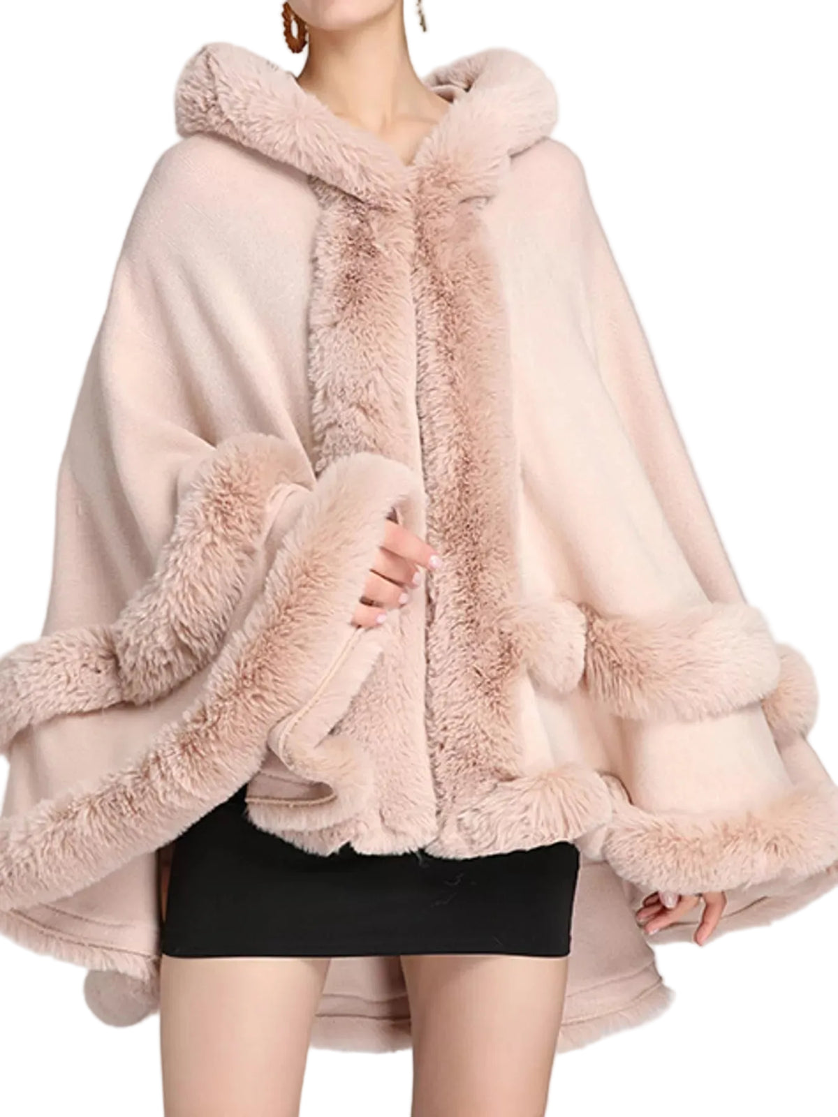 A woman wearing a light pink hooded cape jacket with plush faux fur trim and pom-pom details, paired with a black skirt, against a light gray studio background.