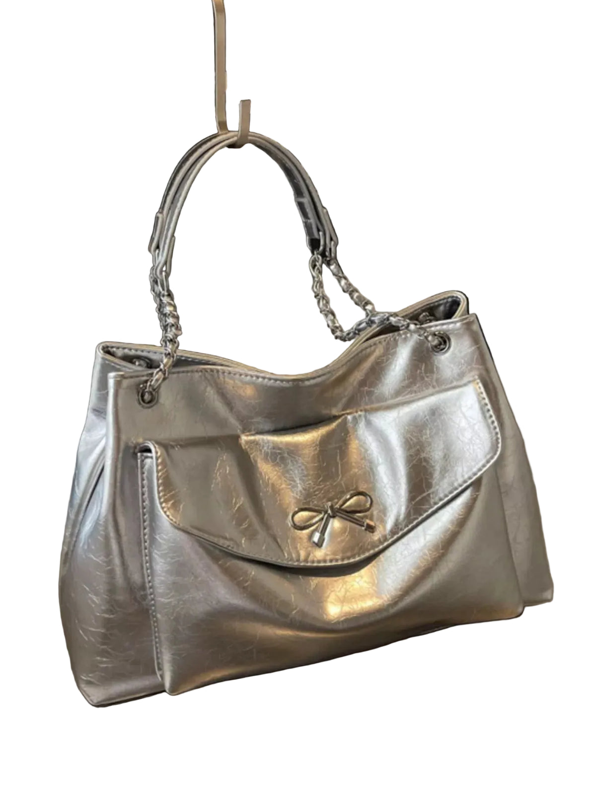 A metallic silver handbag with a front flap pocket and a small bow detail, displayed on a metallic surface with a black framed print in the background.