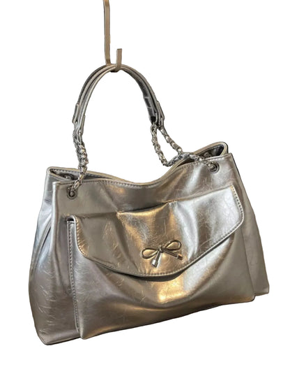 A metallic silver handbag with a front flap pocket and a small bow detail, displayed on a metallic surface with a black framed print in the background.