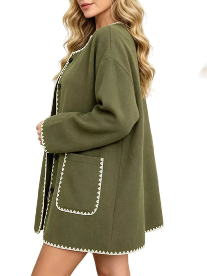 A woman wearing an oversized olive green cardigan with white zigzag trim and black buttons, posed in a cozy living room setting with a sofa and throw blanket.