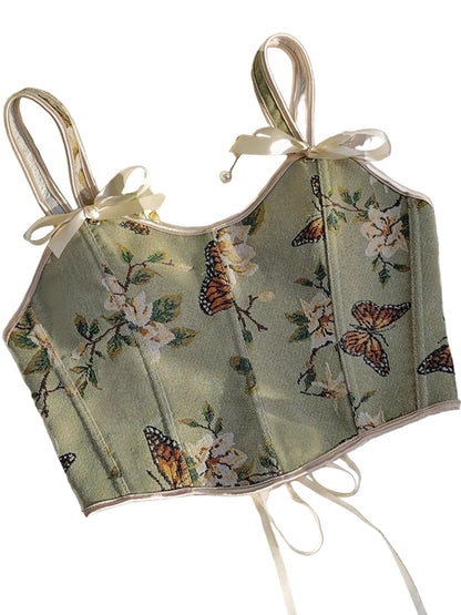 A structured corset-style camisole top with a muted sage green base, featuring a detailed butterfly and floral print in orange, white, and green. The top is styled with cream satin shoulder straps tied in bows, laid flat on a white textured fabric with decorative flowers and a gold mirror frame.