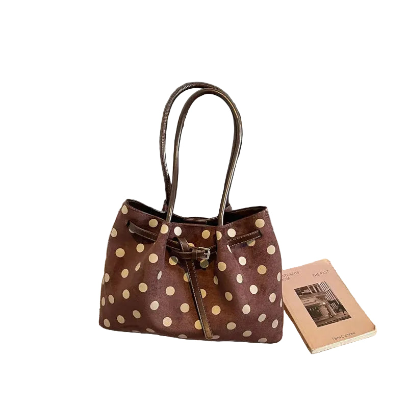 A brown suede-like tote bag with beige polka dots and dark brown leather handles, placed on a textured beige carpet next to a book, with a black bench in the background.