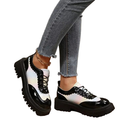 A person wearing black and white lace-up oxford-style platform shoes with distressed gray jeans, standing on a rustic wooden floor.
