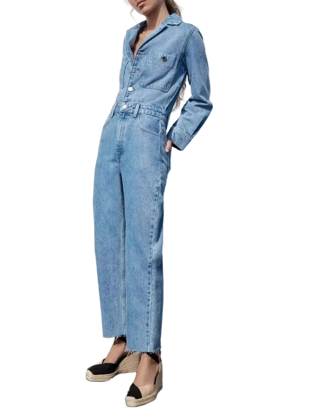 A model wearing a light blue denim jumpsuit with wide legs, standing against a white wall outdoors.