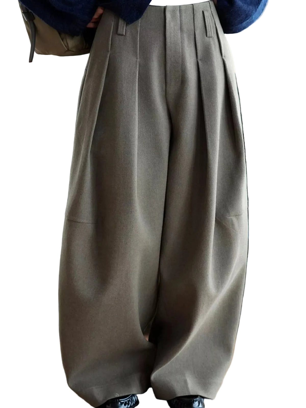 A person wearing very wide-leg pleated trousers in a muted taupe-gray color, standing in front of a black leather sofa on a wooden floor.