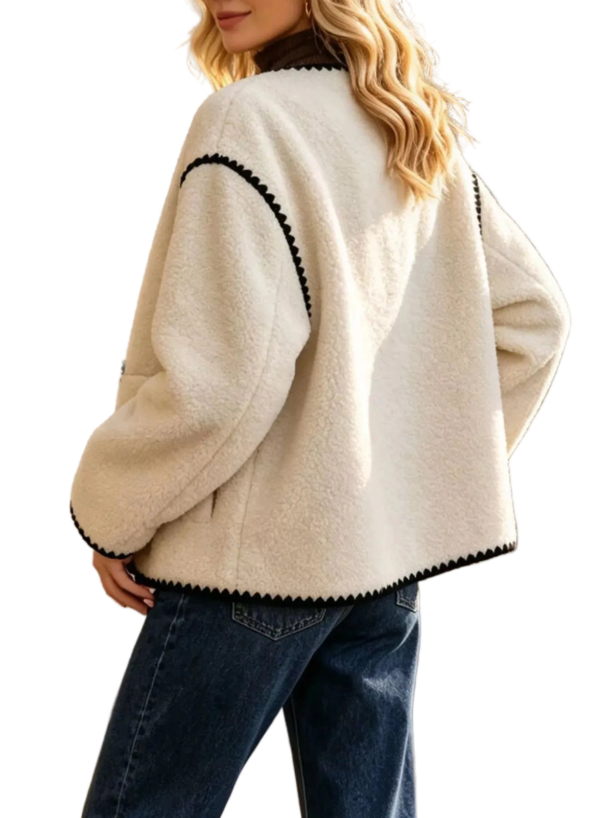 A woman wearing a cream-colored sherpa fleece jacket with black scalloped trim, paired with dark blue jeans, photographed outdoors against a stone wall.